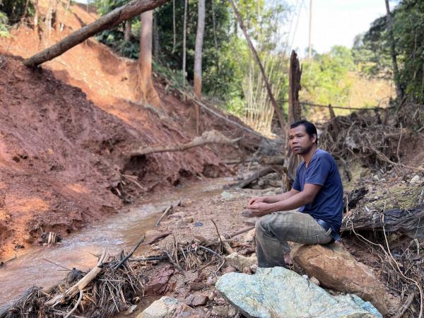 Alai Angah duduk di tebing sungai yang airnya tercemar berwarna merah karat akibat sisa buangan besi.